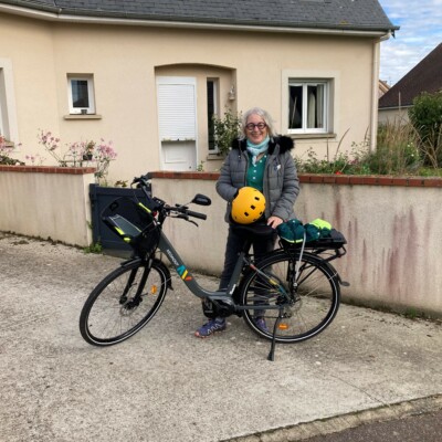 Femme souriante en tenue décontractée et casque, posant avec un vélo devant une maison à Feuguerolles-Bully.